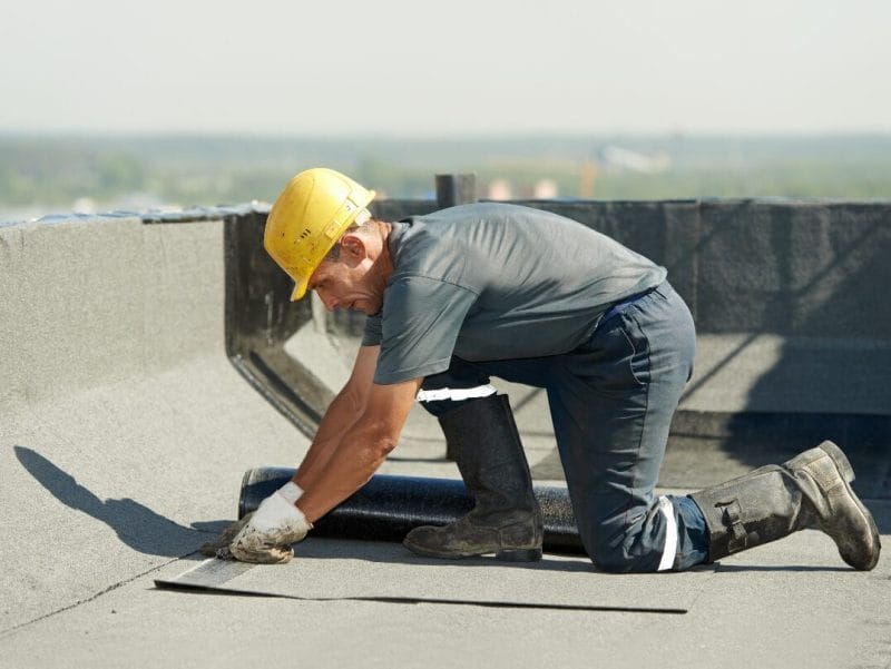 Top 10 Roofing Materials: Pros and Cons 2 A construction worker in a yellow hard hat installing roofing material on a commercial building roof.